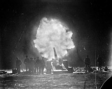 In the black and white photograph, a large artillery gun fires into the night. Personnel are clustered around the artillery, their silhouettes illuminated against the darkened sky. At the muzzle of the artillery, a brilliant white flash bursts forth and contrasts against the black night sky.