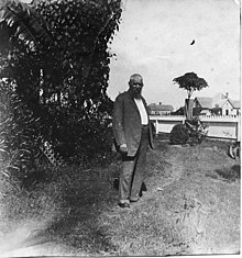 A bearded man standing, wearing a suit. Trees and houses are in the background.