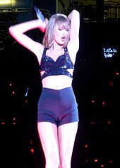 Taylor Swift in a black crop top and shorts, putting her arms on her back, with a microphone on her right arm.