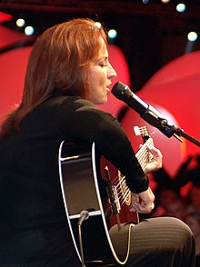 A woman with red hair wearing a black shirt is playing a guitar and facing right