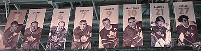 We are looking up at the ceiling of an arena where there are nine banners hanging. On each banner is the picture of a hockey player in a Toronto Maple Leaf uniform. Above each player's photo is a number and the player's name