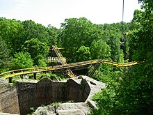 The Loch Ness Monster's second lift hill and mid-course brake run is viewed in the image among green foliage and trees. The mid-course brake run is above the second lift hill in the center image. The tunnel's exterior structure can also be seen in the bottom left.
