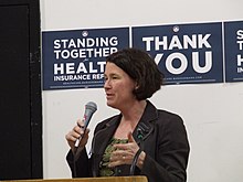 A white woman with medium-length black hair holds and speaks into a microphone before a podium. Behind are signs attached to the wall reading "Standing Together for Health Insurance Reform" and "Thank You".