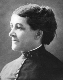 A white woman missionary with her dark hair center-parted and swept up in back; she is wearing a uniform-like plain dark jacket with buttons down the front, and a high collar