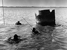 Remains of metal cylinder, 4–6 feet in diameter, protrude from water; three divers in foreground