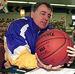An older man wearing a purple and yellow jacket is holding a basketball.