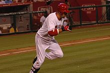 A dark-skinned man with "NGEL" visible in red text on a white jersey runs from home plate to first base. He has on red and black batting gloves, a red batting helmet, white pants and black shinguards.