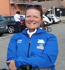 Headshot of a white woman in a bright blue kit with cars in the background.