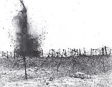 In the monochrome photograph, a shell detonates amidst a field strewn with multiple layers of tangled strands of barbed wire.