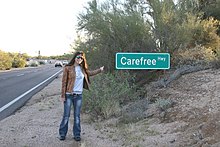Woman standing next to Arizona State Route 74 and pointing to a sign reading 'Carefree Hwy'.