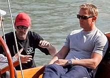 A man wearing a grey shirt, jeans and sunglasses sits on a boat at sea. Besides him, a man wearing a black shirt, red cap, and sunglasses talks to another, who is mostly off the picture.