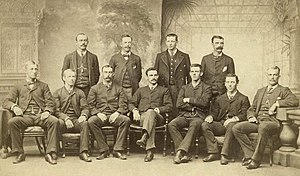 Baseball players are posing for a photograph, four men standing, seven men sitting on chairs.