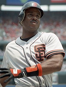 An African-American man looks just right off the camera. His helmet and white jersey both have an orange "S" over the "F" logo on them. The man's left arm is crossed over his body and his right is out of the picture. There is a black and orange glove on his left hand.