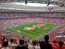 A football pitch is surrounded by a large audience of people mostly dressed in red.