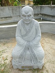 Image of older monk with large eyebrows, sitting, with his hands hidden in his sleeves