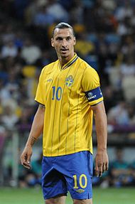 A photograph of a man with dark hair wearing a yellow football shirt, blue shorts and a dark blue captain's armband on his arm, the man is looking away from the camera.