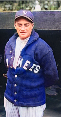 A man in a white baseball uniform and a navy blue sweater and cap