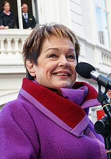 A woman (Nørby) in a purple coat at a microphone
