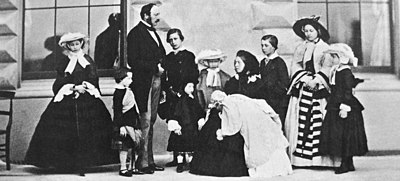 Victoria, dressed in black, is seated and holding her infant daughter. Prince Albert and their other children stand around her.