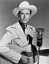 A young man wearing a cowboy hat, a jacket and tie, playing a guitar at a microphone