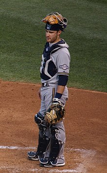 A man wearing a gray Brewers jersey, gray pants, navy blue cap, and catchers gear standing at home plate