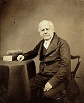 A middle-aged man in Victorian clothes, seated.