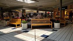 The concourse of a shopping mall with white and brown tiled floors. Several storefronts are visible, as are several brown wooden benches surrounding a circular fountain