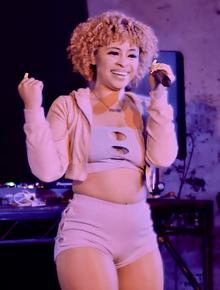 Ice Spice, dressed in pink, smiles and performs with a mic in her hand.