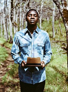 Tunde Baiyewu standing outdoors, wearing light blue denim shirt and holding a brimmed hat, looking directly at camera