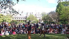 An outdoor crowd watches two performers. Behind them is an attractive white building with spires and a colonnade.