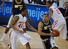 A basketball player in a dark blue uniform drives with the basketball and is defended by a player in a white uniform.