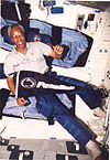 man wearing polo shirt and cargo pants, holding Penn State pennant, background interior of Space Shuttle