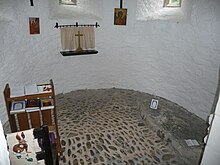 The interior of the apse has a small shelf with books, a crucifix, and a large stone slab on the floor, indicating Melangell's grave.