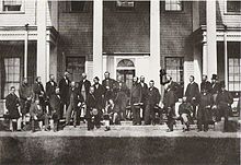 A black and white photograph of several men, standing and seated outside of a row of buildings