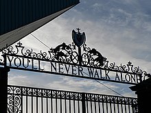 Design of the top of a set of gates, with the sky visible. The inscription on the gates reads "You'll Never Walk Alone".