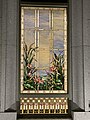 A stained glass window from the Orem Utah Temple, depicting bullrushes and cherry blossoms, next to water elements, with a blue sky with clouds. It is framed by a golden rectangular frame, and has stone masonry surrounding the window.