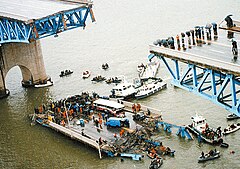 The picture is taken from a vantage point above the bridge disaster. The middle section of the bridge is missing, and spectators with umbrellas can be seen observing the collapsed section of the bridge which is floating in the water. Multiple small boats and kayaks can be seen crowding around the collapsed section of the bridge. There is a large crowd of people seen standing on the collapsed section of the bridge, along with a severely damaged bus, van, and car.