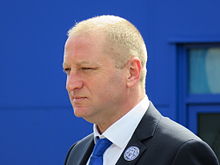 A man with thinning blonde hair and narrowed eyes, wearing a dark jacket, white shirt and blue tie looking straight ahead