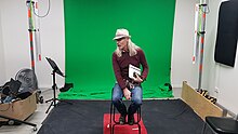 Waka sits on a stool in front of a small green screen. Film equipment and boxes are at the edges of the room. Waka has a hat on and in his hand is a folder.