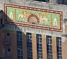 Closeup of a portion of a building, with a multicolored relief decorated with a sunburst, mythological animals, and beehives, on a green background. Below the relief are long piers of windows and brick walls.