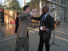 A white man, Gavin Newsom, wearing a gray suit reaches to embrace Jones, while holding a book in his right hand. Jones, who is also reaching out, wears a dark suit and has a microphone and piece of paper in his left hand. Inside a glass-walled building behind them, a display says "Climate is an angry beast and we are poking at it with sticks".