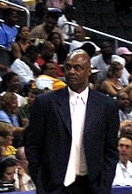 A man, wearing a black suit, white shirt and white tie, is standing in front of a crowd.