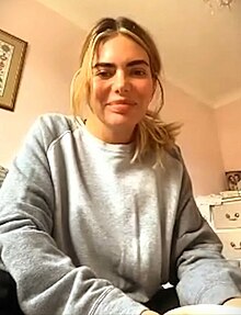 A grainy image of a blonde woman in a room of a domestic house wearing a gray jumper.