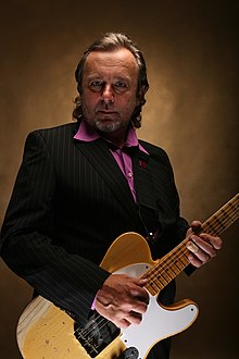 Michal Pavlíček wearing a dark pin-striped suit and purple shirt, holding an electric guitar, looking intensely at camera