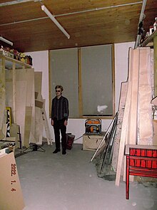 A blonde haired man in all black with sunglasses standing still in the corner of a studio, alone