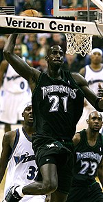 A black basketball player wearing a green jersey prepares to dunk a basketball