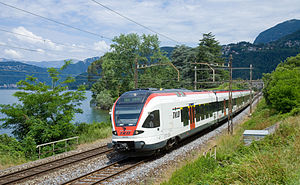 White-and-red train on tracks next to lake with mountains in the background