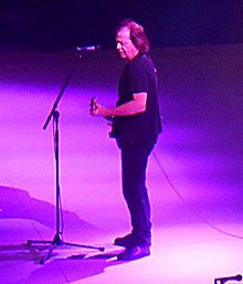 Stevie, 59 years old, shown in full left profile. Strums guitar while at microphone on a stand. He wears black T-shirt, dark blue pants and grey-blue runners.