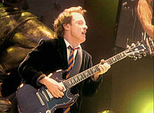 Angus, aged about 45, is shown in an upper body shot wearing his trademark schoolboy uniform. He faces away to his left, with his left hand halfway down the neck of his guitar.