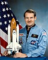 man in flight suit holding Space Shuttle mode, American flag in background